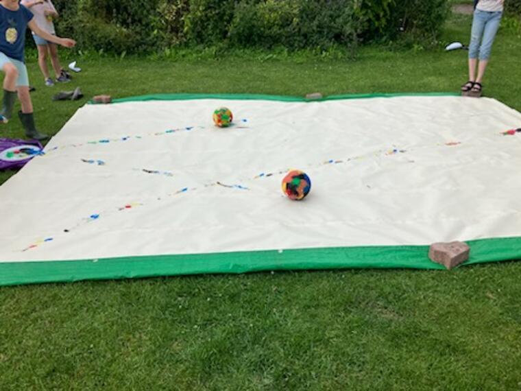 Ein großes Papierbild liegt auf einer Wiese. Kinder schießen Farbe mit einem Fußball darüber.