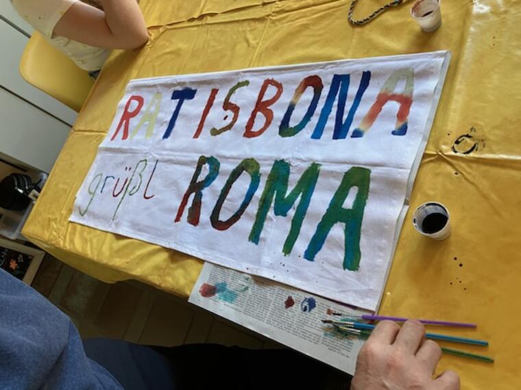 Ein Stoffbanner, auf dem mit Stofffarben gerade der Schriftzug "Ratisbona grüßt Roma"aufgemalt wird